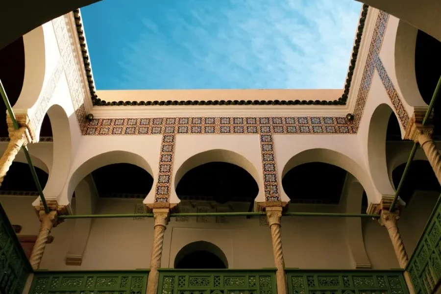 Interior courtyard with white arches, carved columns, and green decorative railing, framed by patterned tile bands and a blue sky overhead viewed from below.