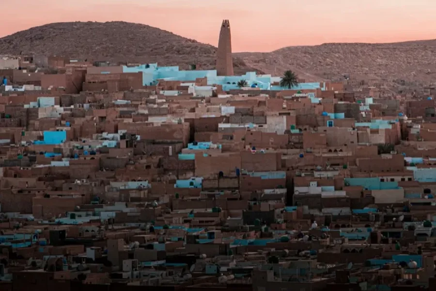 Sunset over a dense desert town of brown adobe houses with blue accents and a tall stone tower in the background.