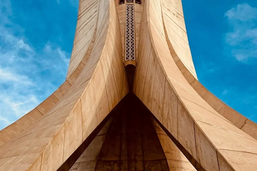 Low-angle view of a beige stone monument with sweeping arches converging at a central point against a blue sky.