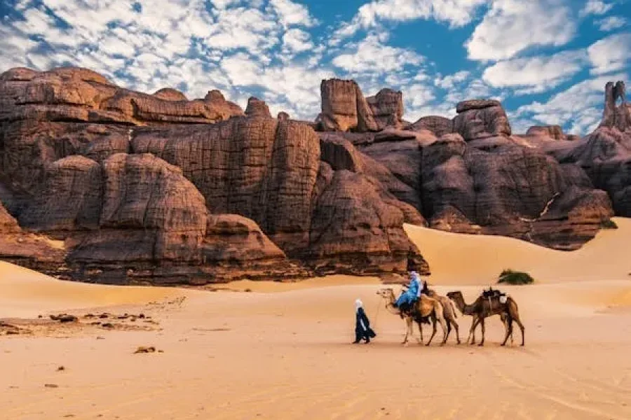 Traveler leading two camels across a golden desert with dramatic red rock formations and a blue, cloud-filled sky.